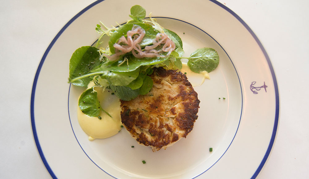 Overhead view of crab cake and salad on a white plate decorated with a small blue anchor