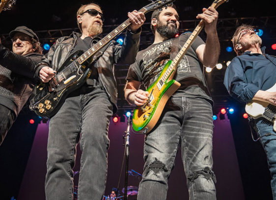 Four men standing close together and playing guitars onstage