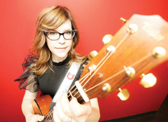 Woman playing a guitar against a red background, and her guitar is so close to the camera that it is out of focus