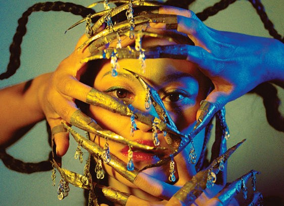 Woman with her hands in front of her face, painted blue and gold, with long decorative fingernails attached