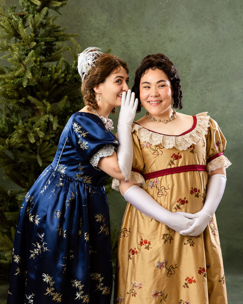 Two women in Regency attire, one whispering into the other’s ear